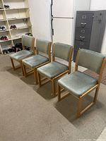 Photo showing four oak frame dining chairs with teal velour upholstery on seats and backs, arranged in a row on a beige carpeted floor in a room with shelving, a refrigerator, and a filing cabinet.