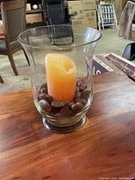 Transparent glass vase on wooden table containing an orange pillar candle and smooth brown stones at the base.