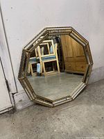 Photo shows a 10-sided ornamental mirror with a gold beveled frame resting against a wall, cardboard and wood furniture in background.