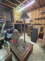 Two metal lamps in a workshop setting: one taller floor standing lamp with a conical metal shade and tripod base, and one smaller table lamp with similar metal shade visible in the background on top of furniture.