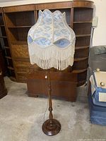 Floor standing lamp with wooden base and decorative fabric shade featuring blue floral prints and fringed trim.