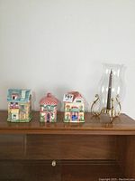 Front view of all three ceramic biscuit jars and the glass candle holder with chimney on display top of wood cabinet.