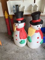 Pair of large plastic blow mold snowmen with black top hats and red scarves, each holding a yellow broom, alongside tall red plastic candle decorations.
