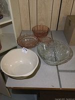 Photo showing five bowls: one large 12-inch stoneware mixing bowl in cream and yellow, one large clear glass bowl with floral pressed pattern, and three pink depression glass bowls, two footed.