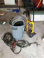 Overview of lot showing yellow hand truck, gray trash can with coiled hose, welding cables on floor.