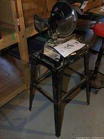 Jobmate compound miter saw on metal base with user manual on top, seen from the side showing the entire unit and stand.
