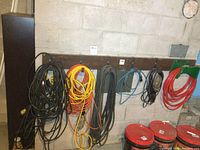 Wide shot showing multiple extension cords of various colors hung on a wooden rack against a concrete wall. Also visible are coiled air hoses and red buckets on the floor.
