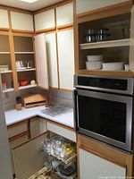 Kitchen corner showing open upper and lower cabinets fitted with various glass and ceramic bakeware and containers, wood bread box, and built-in oven visible.