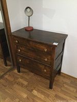 Side angled view of solid wood dresser with vintage lamp on top showing three drawers and brass knobs.