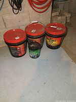 Three large pails of Airport Grade Asphalt Driveway Sealer and one smaller can of waterproof sealant placed on the floor.