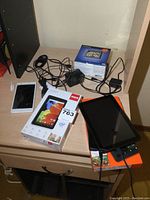 Overview photo of electronics including tablets, GPS in box, phone with cracked screen, and chargers on wooden surface.