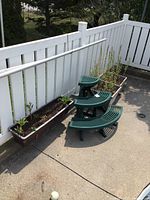 Green resin corner plant stand with three tiered curved platforms next to three long rectangular brown resin planters outdoors on concrete patio with white fence background.