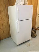 White Frigidaire refrigerator with top freezer, showing side and front view against wood panel wall.