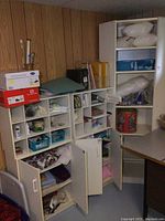 Wide view of multiple white cubical shelves and tall cabinet with doors open revealing contents including fabric, craft baskets, tins, and fiberfill bags