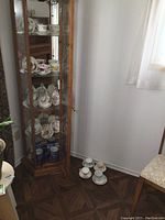 Photo of wooden curio cabinet with shelves holding various tea cups and saucers, some pink and yellow floral designs visible, and coffee mugs placed on floor beside curio.