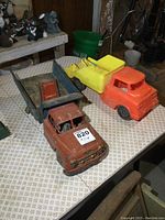 Two vintage trucks on a patterned table: front view shows orange Lumar steel dump truck with blue bed and red interior bed, and bright orange plastic dump truck with yellow bed