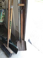 Full view of several yard tools leaning against wall: a square blade shovel, a broom with wooden handle and black bristles, and a floor squeegee with a green handle. Shows metal and wooden components with wear.