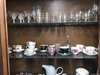 Overall view of glass shelves inside wooden cabinet showing a variety of glassware on top shelf and a row of porcelain china teacups and saucers on the center shelf.
