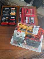 Photo showing three bit sets: Craftsman drill and countersinking bit sets in black and red plastic cases, and Dewalt screwdriving bit set in sealed blister packaging on a wooden surface.