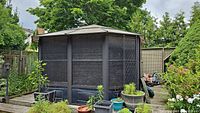 Photo of a 12x12 gazebo with mesh walls, dark metal framing, surrounded by garden plants.