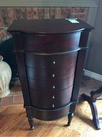 Front view of the dark wood jewelry cabinet showing the curved front, 5 drawers with metal knobs, and floral carving detail on the upper drawer.