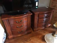 Pair of wooden nightstand chests of drawers shown side by side with flat tops and three drawers each, ornate metal handles, warm brown finish, placed under a TV with minor surface wear visible.