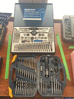 Open metal case showing the full tap and die set with drill bits, dies, taps, wrenches, and screwdriver parts organized in slots.
