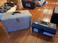 Photo showing two tool boxes on wooden floor, one large metal diamond plate style with latch and black handle, one smaller black plastic with stickers and handle.