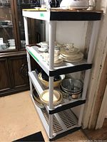 Full view of white plastic utility shelf with four tiers containing dishes and kitchenware items in basement.