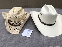Two kids' medium-sized Modestone cowboy hats placed side by side on a textile surface, one white with a studded black band, one off-white crocheted straw with a brown band.