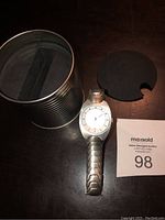 Photo showing the silver Cross wristwatch along with its original cylindrical tin packaging missing the top lid and a foam insert.