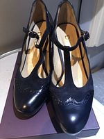 Close-up image showing navy blue women's T-strap shoes on box, with suede and glossy leather textures and decorative wingtip edges on toe area.