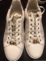 Front view of white and beige Michael Kors sneakers with MK logo charm on laces, showing toe caps and lacing detail.
