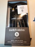 Front view of boxed Switch Sticks walking stick in black with visible wooden handle and branding label on the box front.