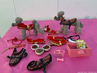 Two grey felt poodles standing on pink surface surrounded by various dog outfits, collars, food bowls, a pink pet box with a box of dog food, a pink bone, and two plaid harness items.