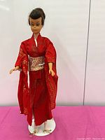 Full view of the vintage brunette ponytail Barbie doll standing dressed in a red kimono with gold leaf patterns, holding a white samisen accessory. Doll is posed on a stand (stand not included in sale).