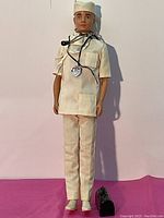Front view of Ken doll standing on pink surface wearing cloth medical scrubs and cap with stethoscope draped around neck, black medical bag placed by feet