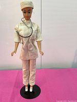 Full frontal view of Barbie doll wearing white doctor outfit with cap and stethoscope around neck, standing on a black stand.