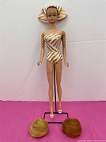 Full doll standing on metal stand wearing gold and white striped swimsuit and matching headscarf, two wigs and shoes shown on ground