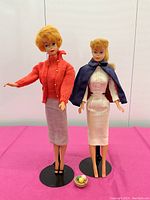 Front view of two vintage Barbie dolls, one in red sweater and pencil skirt, the other in white dress with blue cape, posed on stands with a small basket of knitting accessories at their feet.