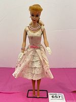 Front view of the Barbie doll wearing original 1960s pink polka dot dress with lace trim, pearl jewelry, white gloves, and black display stand on pink background.