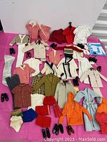 Photo showing a wide assortment of Ken doll clothes and accessories including sweaters, coats, shoes, and bathing suits laid out on pink cloth.