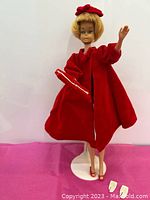 Barbie doll wearing red coat and hat posed on stand with red and white shoes visible