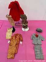 Photo of the vintage Barbie outfits displayed on stands and laid out flat showing dresses, suits, coat, and accessories on pink background.