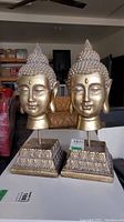 Two gold-toned Buddha head sculptures side by side on decorative stands shown in ambient lighting.