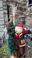 Wide shot of the snowman holding skis next to the artificial Christmas tree and reindeer figure with stone wall background.