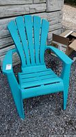 Front view of one turquoise molded plastic Muskoka chair showing slatted back and seat, wide armrests, placed outdoors on gravel near wall and boxes.
