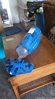 Blue Pool Blaster handheld pool vacuum cleaner placed on a wooden table, showing front transparent suction nozzle
