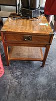 Full view of the wood side table showing drawer, rattan bottom shelf, and brass corners