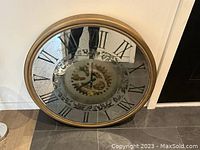 Photo showing the large 32-inch round wall clock with a gold frame and Roman numerals, leaning against a wall on a tile floor.